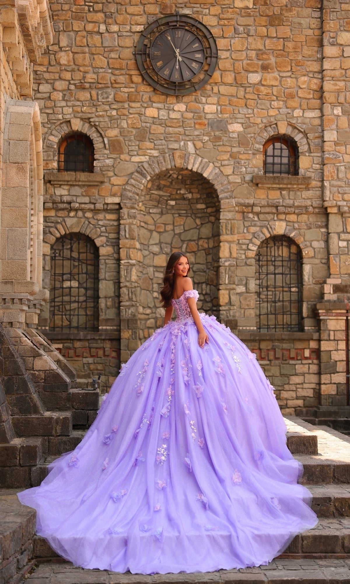 A woman in the Amarra 54231 3D Floral Quinceañera Dress, featuring large lavender flowers, stands on stone steps before an old building with arched windows and a big clock.